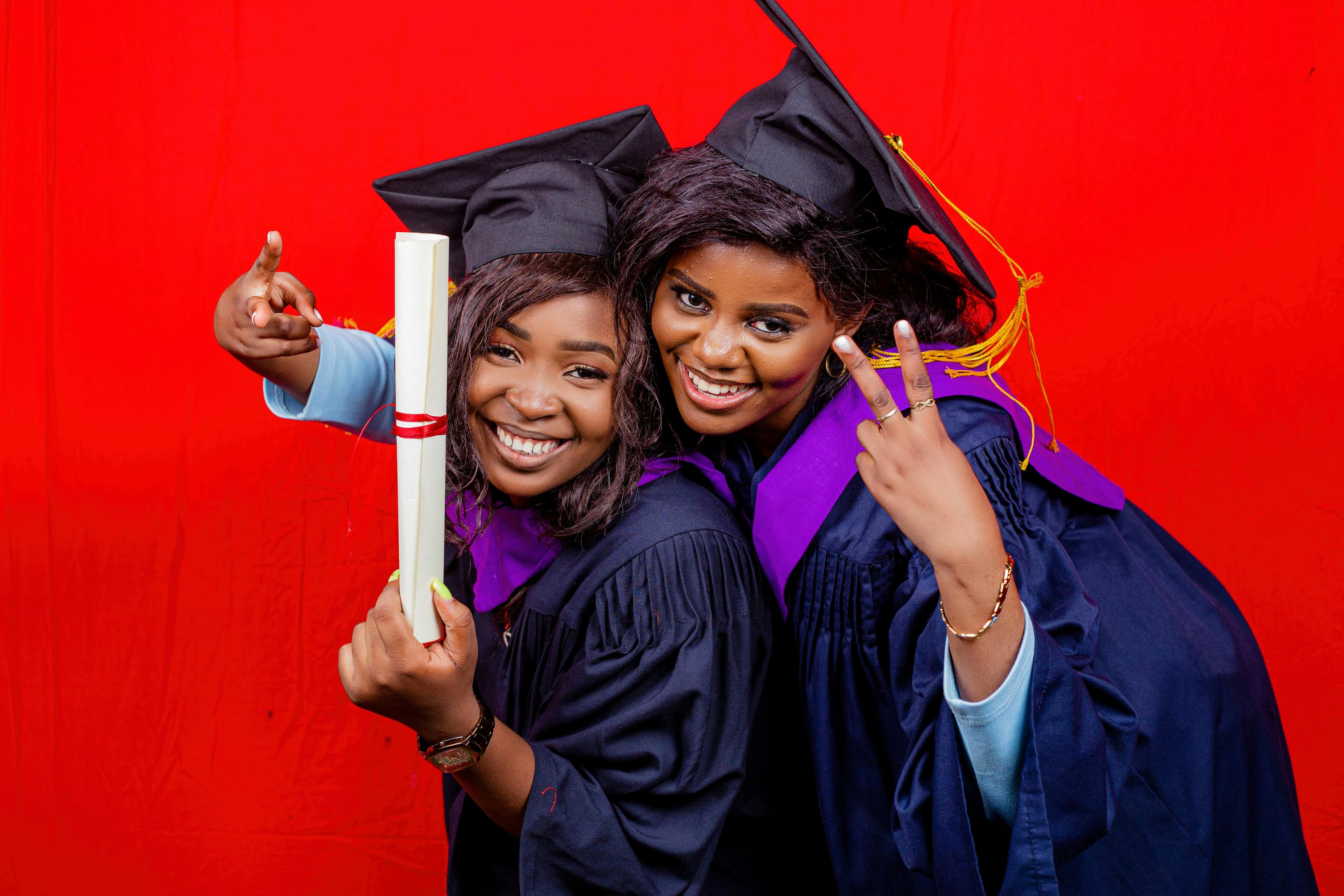 Two graduates in caps and gowns celebrating with diplomas and smiles against a vibrant red background.