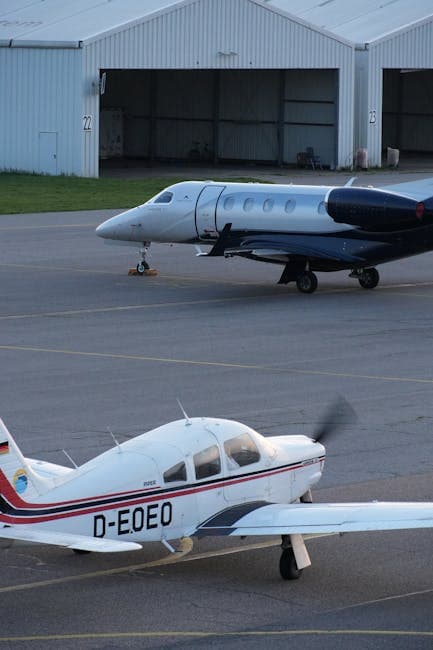 Two private aircraft stationed at a European airfield with hangars in the background.