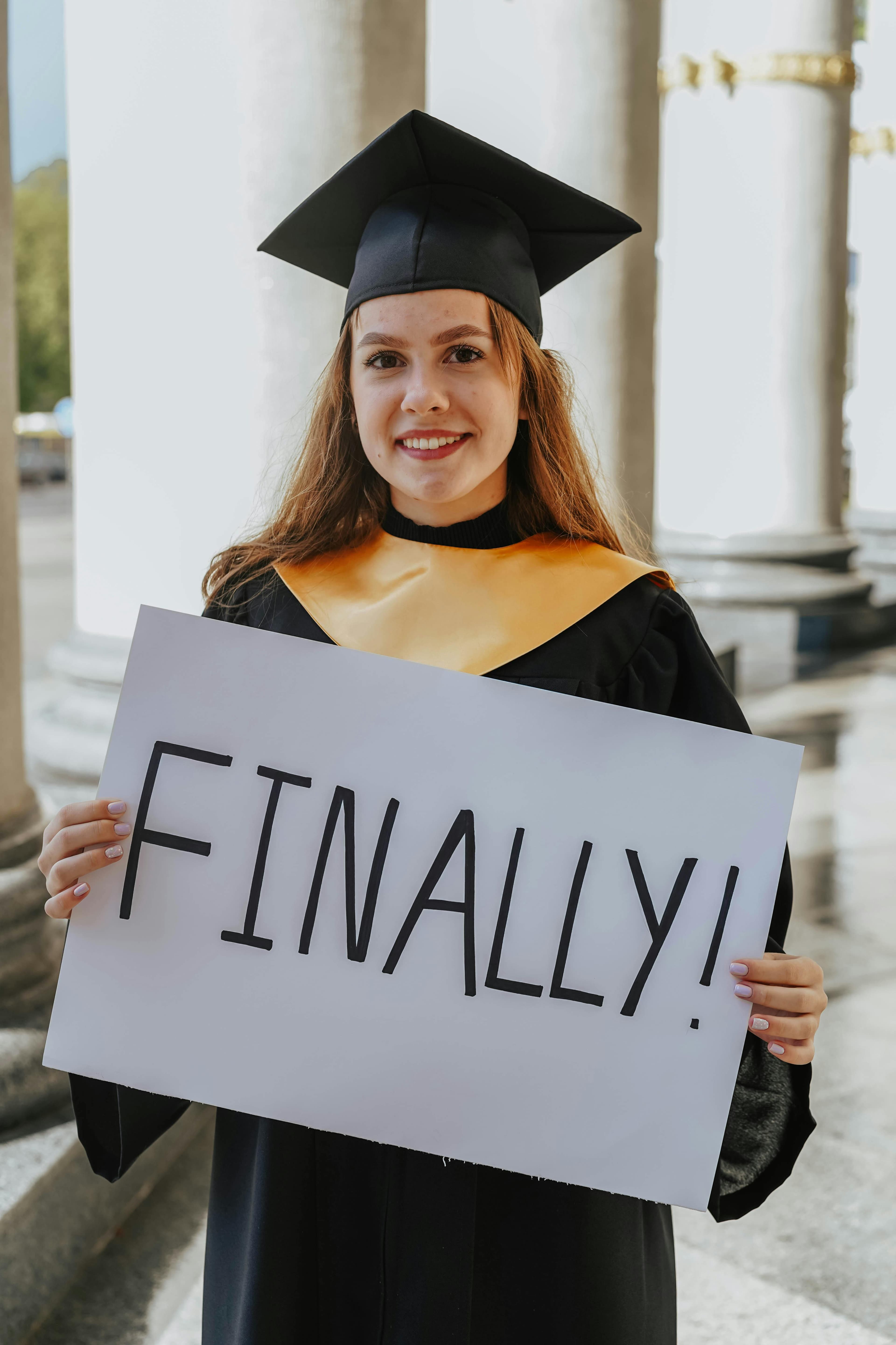 A joyful young woman in a graduation gown holding a sign that reads 'Finally!' outdoors.