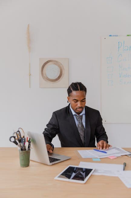 Businessman with braided hair working on documents in an office setting with a laptop and paperwork.