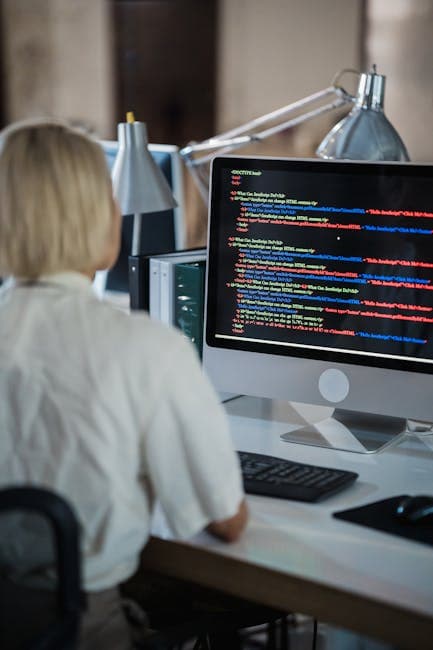 Professional woman coding at office desk with two monitors and desk lamps.