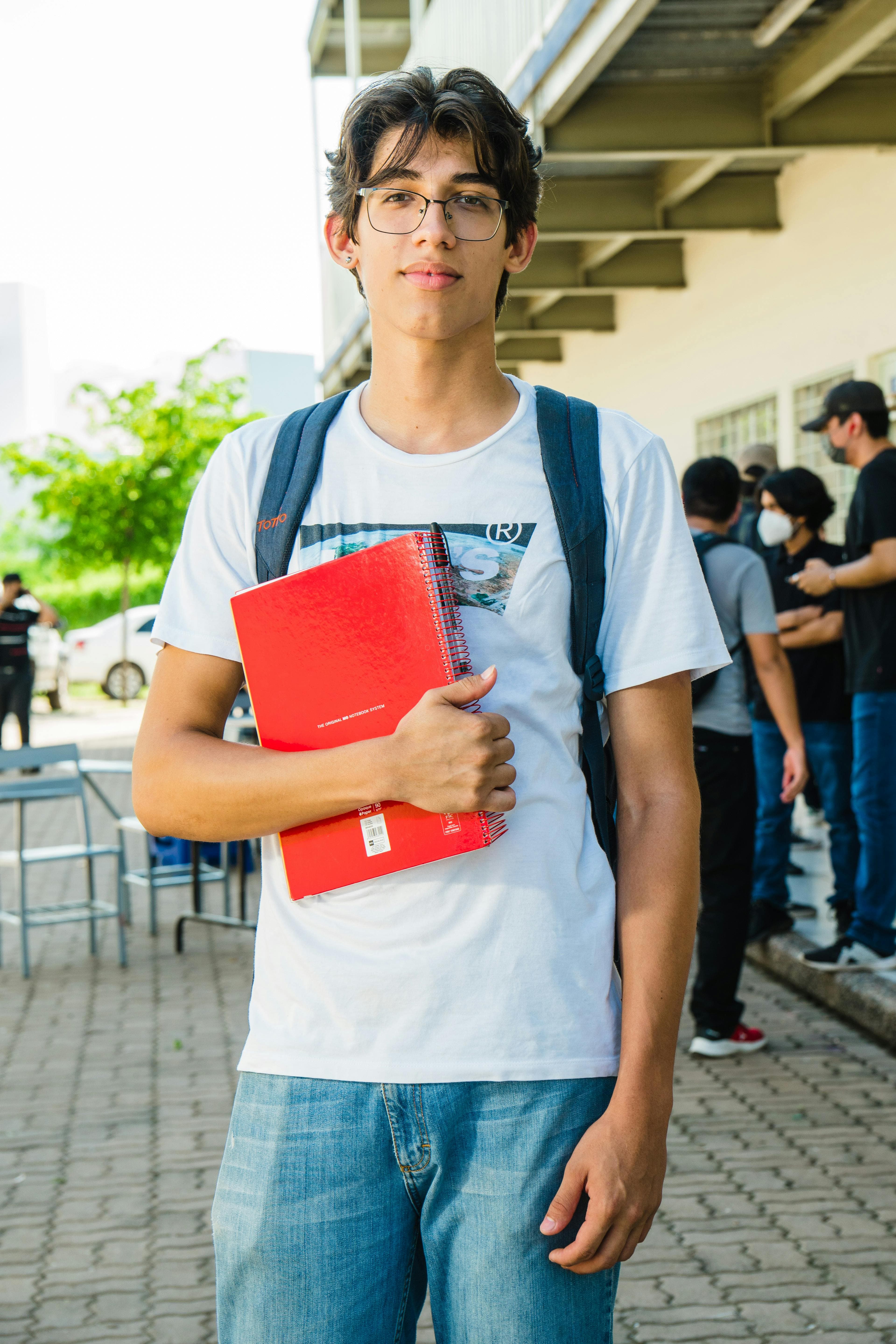 Hispanic university student outdoors in Culiacán holding a red notebook, wearing casual attire.