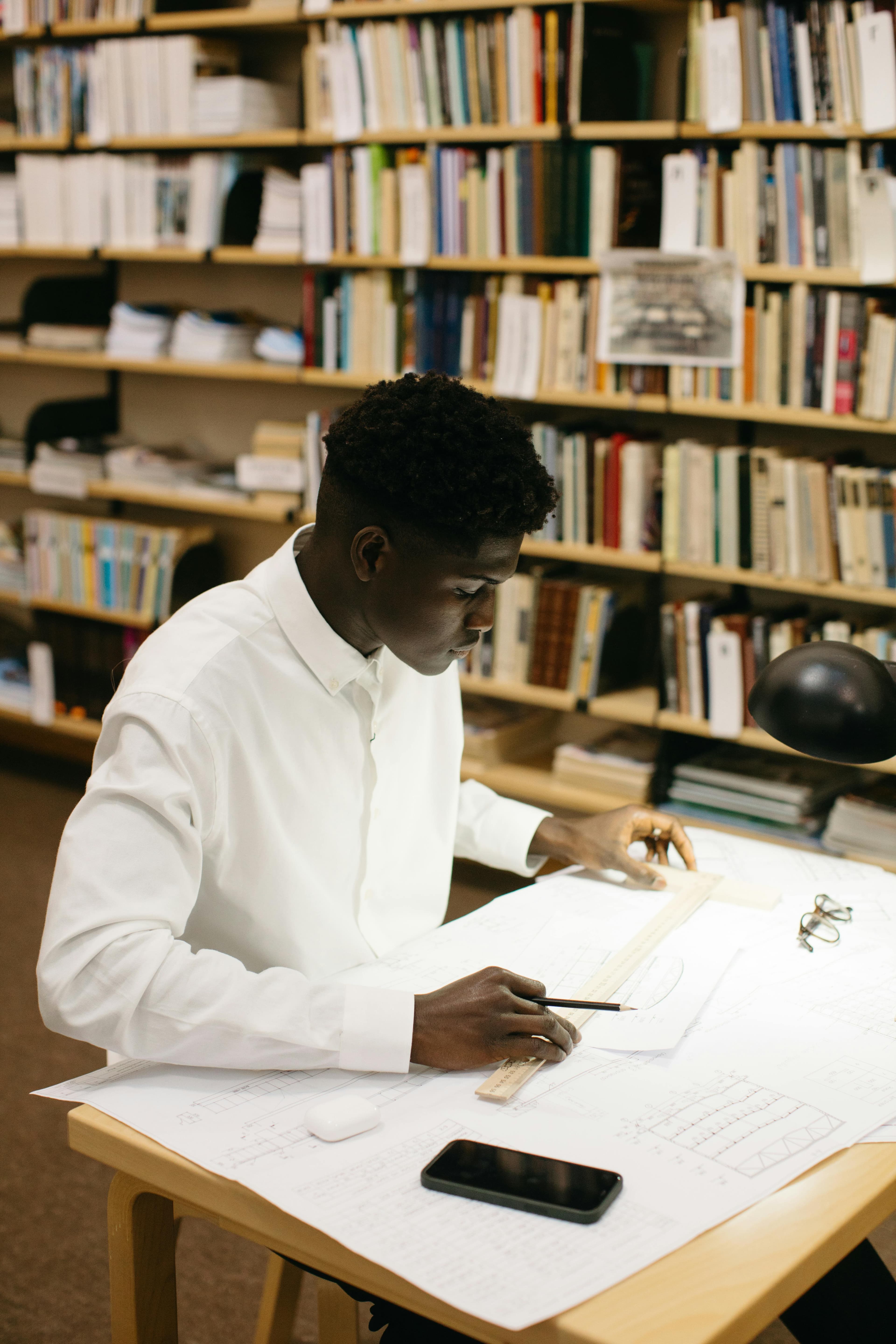 شاب في مكتبة يعمل على تصميم على مكتب بتركيز واهتمام. مثالي للموضوعات التعليمية والإبداعية.